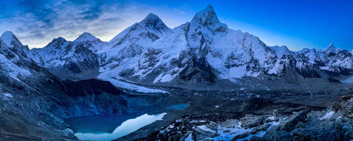 A panoramic fine art photograph captured near Kala Patthar overlooking the Everest region of Nepal. The composition features Mount Everest, Nuptse, surrounding Himalayan peaks, glacial lakes, and rugged moraines bathed in the cool light of dawn, conveying the immense scale and timeless beauty of the world's highest mountain landscape.