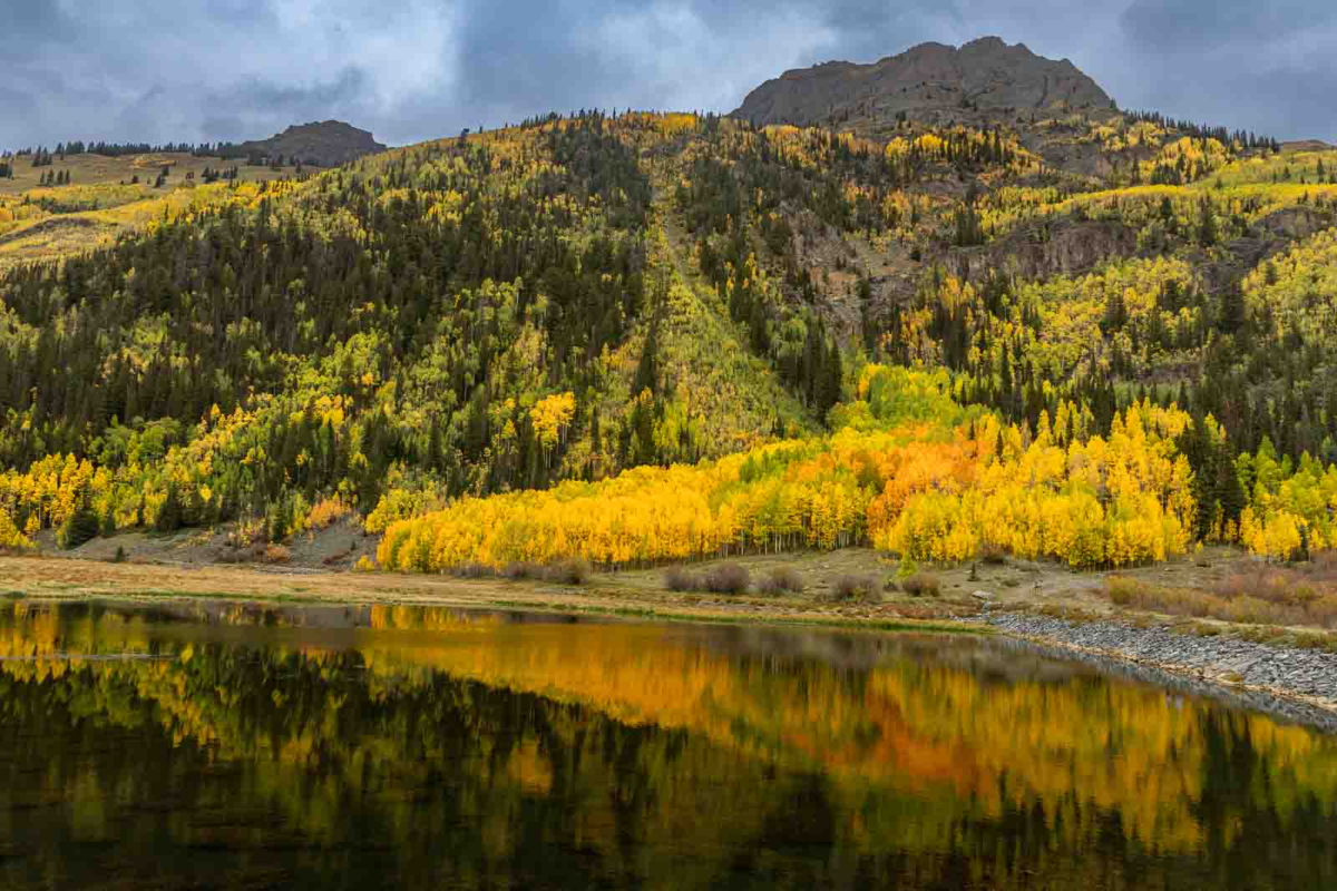 Golden aspens and a mountain ridge reflected in Crystal Lake beneath a cloudy autumn sky