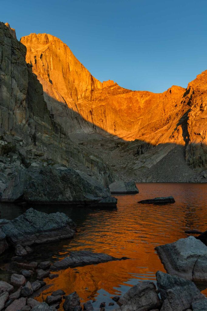 Golden sunrise illuminates Longs Peak’s east face above Chasm Lake, with orange reflections and rocky foreground in Rocky Mountain National Park.