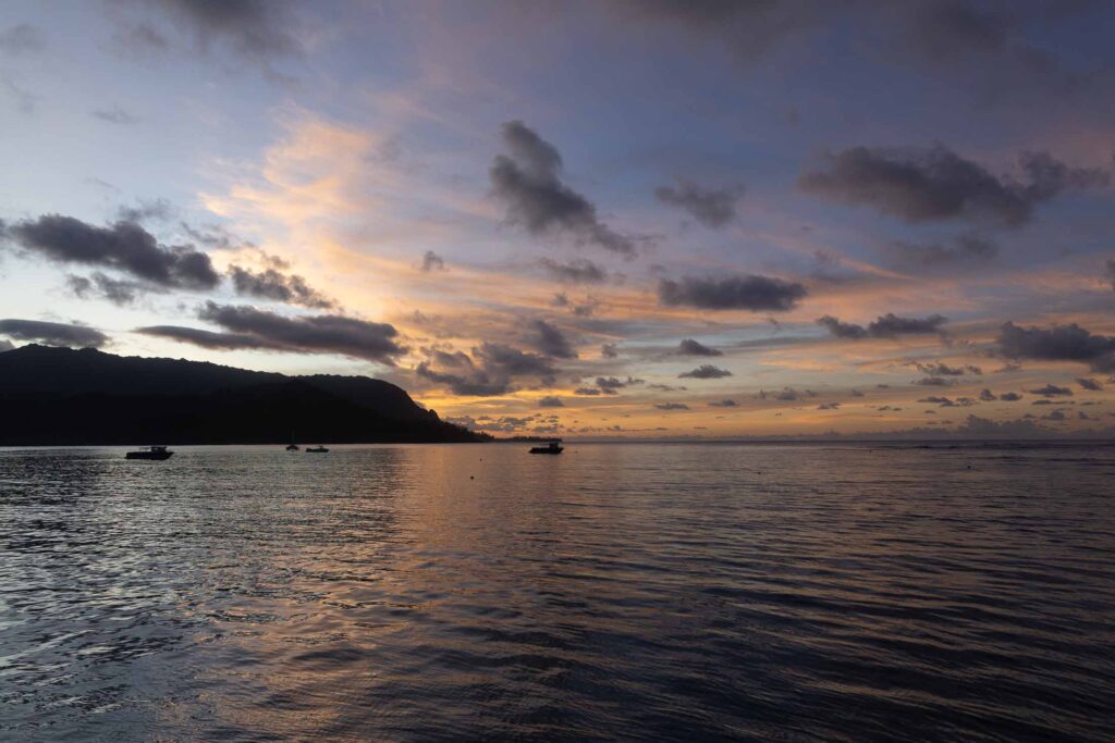 Anchored boats on Hanalei Bay beneath orange sunset clouds and a dark mountain silhouette.