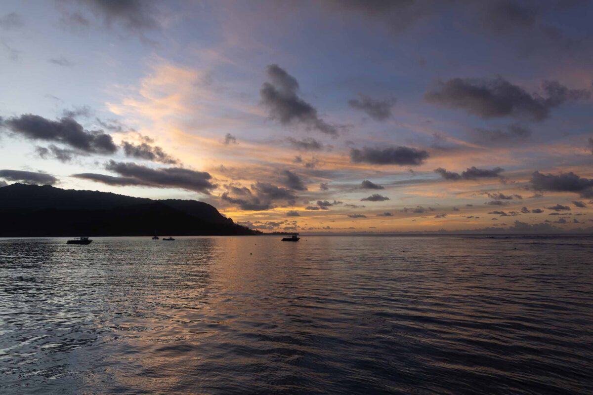Anchored boats on Hanalei Bay beneath orange sunset clouds and a dark mountain silhouette.