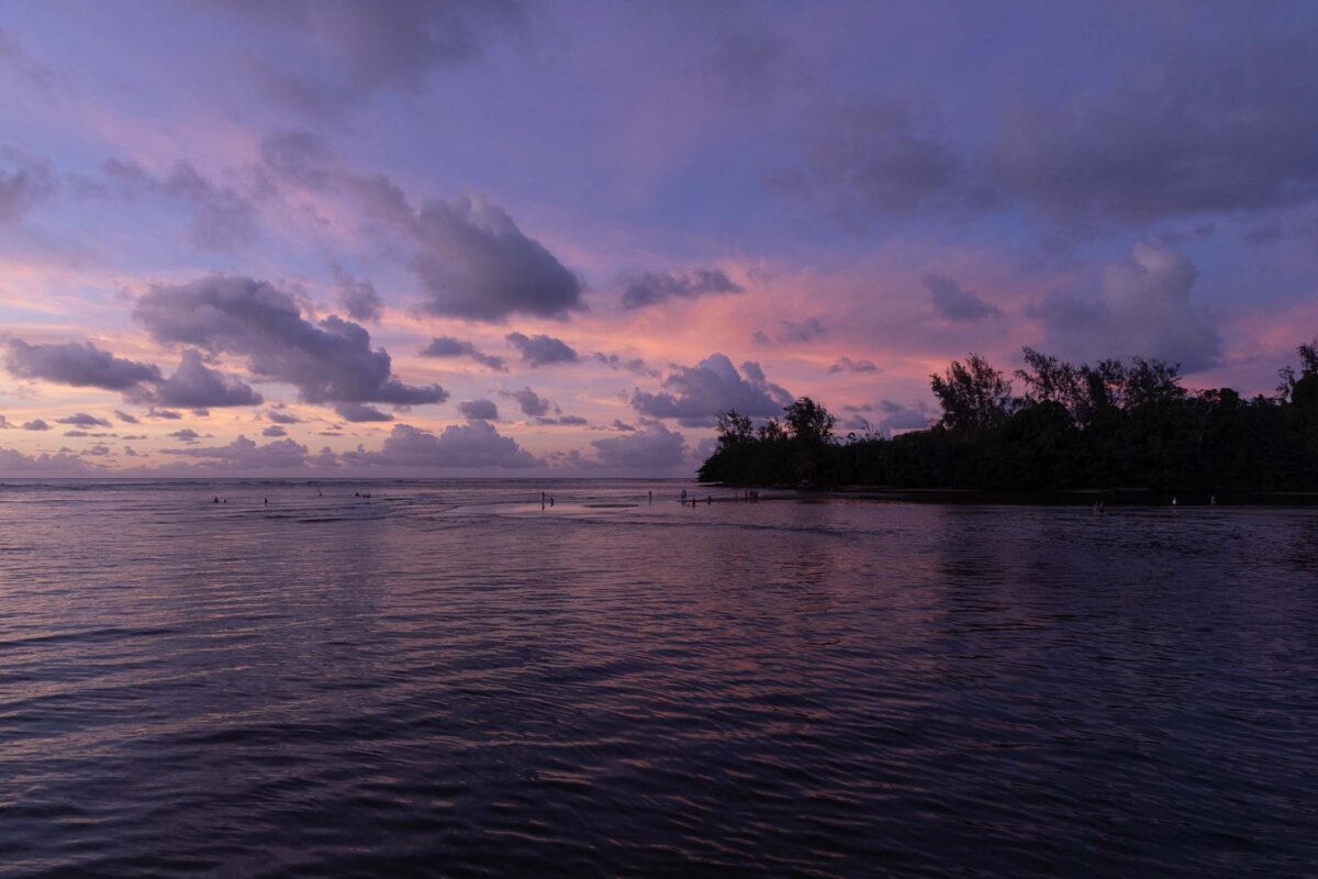 Pink and purple sunset clouds reflected across Hanalei Bay, with a dark wooded headland on the right.