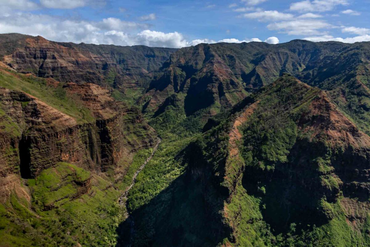 Aerial view into Waimea Canyon with layered red rock walls, green ridges, and a winding river below.