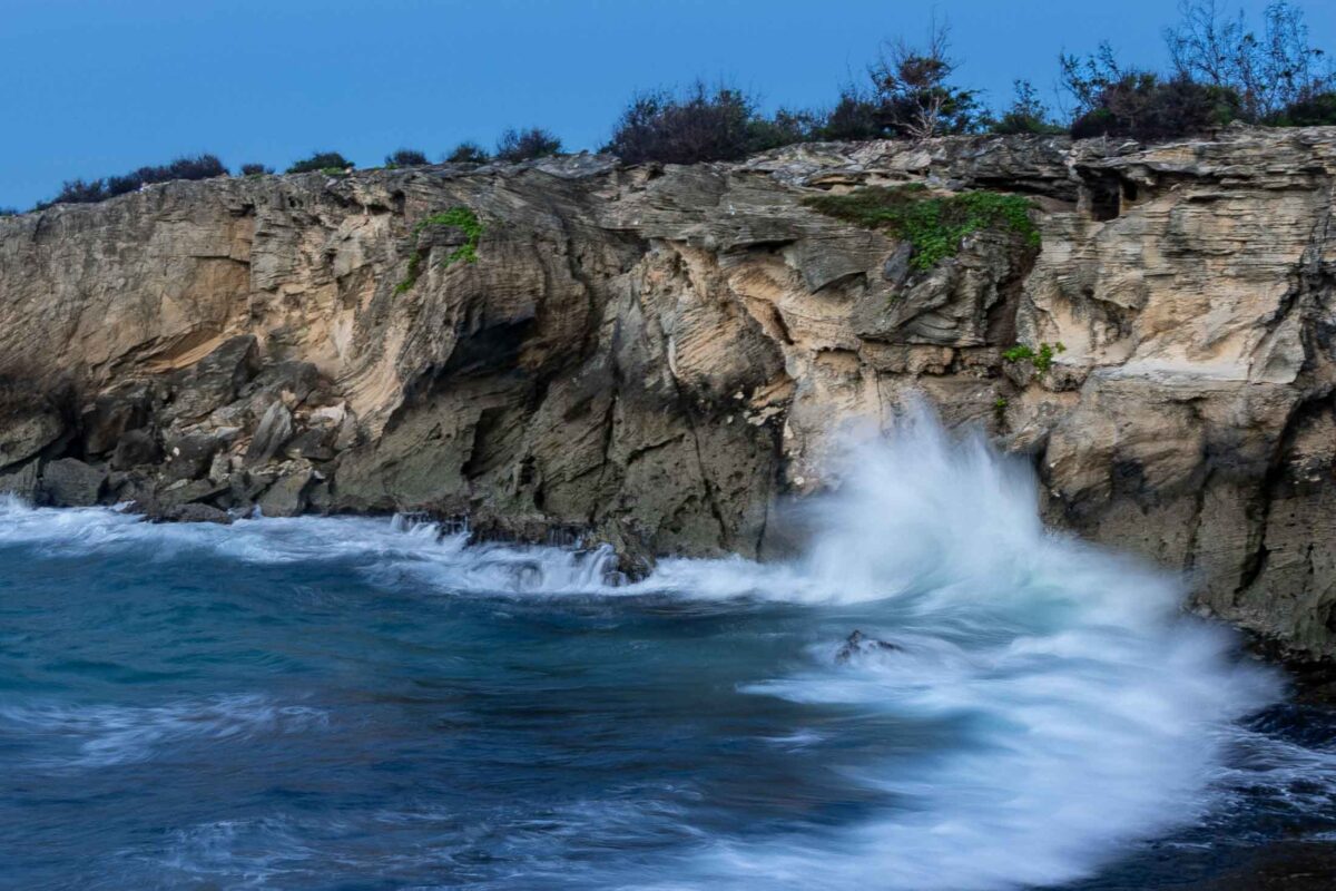 A breaking wave sends white spray against textured coastal cliffs at Shipwreck Beach, Kauai.